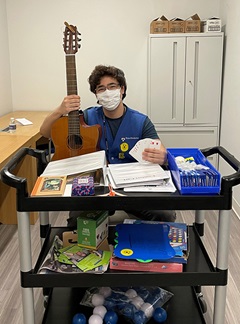 Hospital volunteer Yihan Xie, holding a fanned-out deck of cards in one hand and a guitar in the other, sits behind the HUP Comfort Cart, which is stocked with games and other freebies.